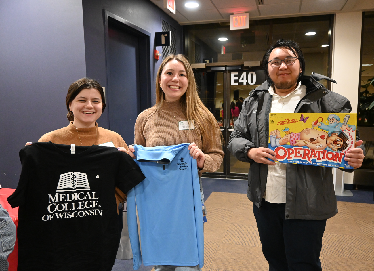 group of 3 students holding MCW shirt, WisMed sweater, and Operation game